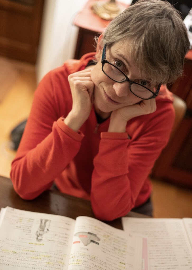 A white woman with shrt grey hair wearing black-framed glasses and a red shirt leans her elbows on a table where a scientific textbook is lying open.
