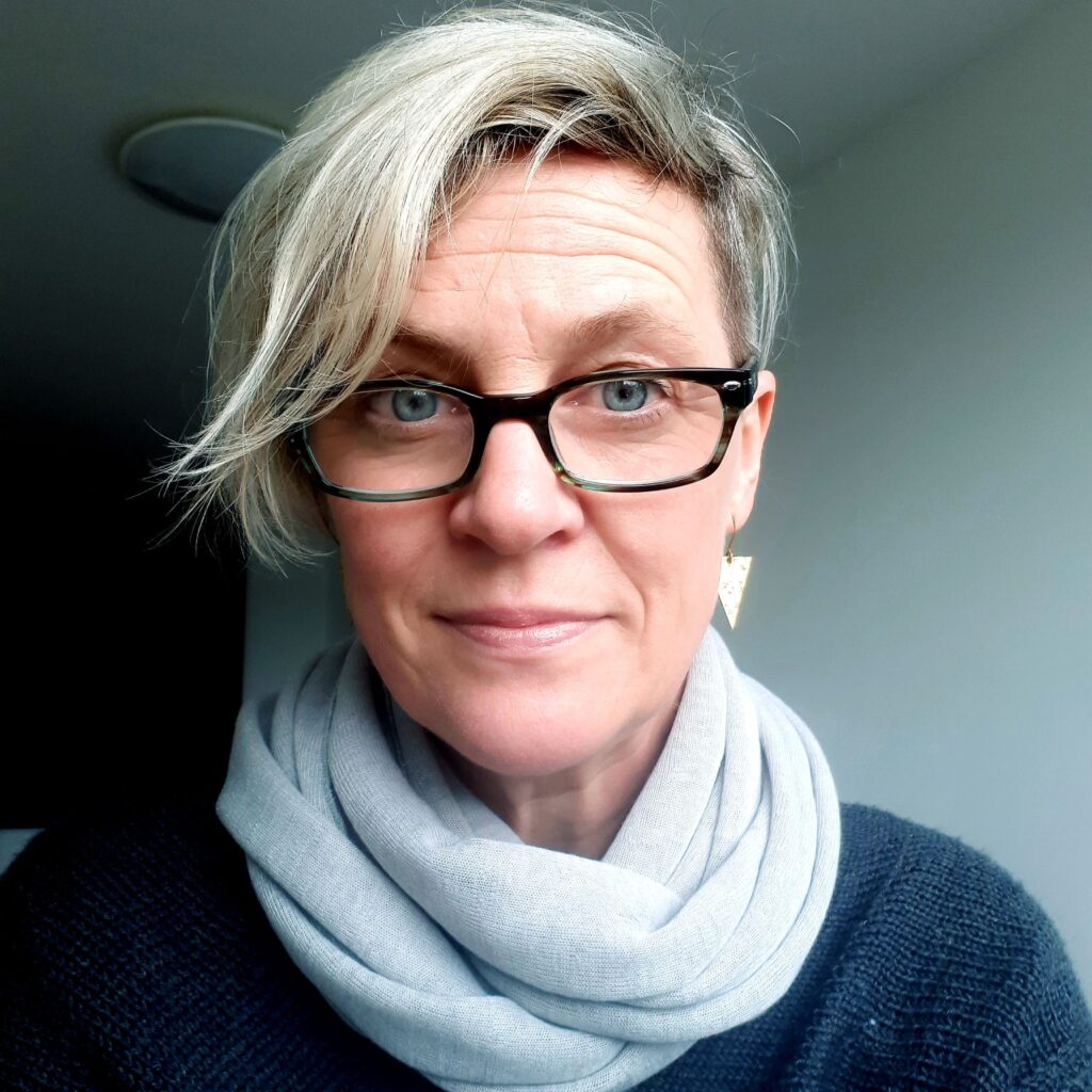 Jo Bell is a white woman in her fifties with silver-grey hair cropped short at the sides and swept over in a flowing quiff. She is wearing large rectangular dark tortoiseshell glasses, and a pale blue scarf looped over a dark blue jumper. She looks mildly surprised.