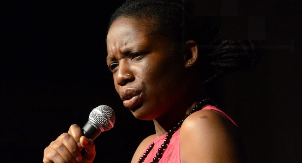 Kat is a black woman in her late forties. She is looking away from camera, to the left of frame, and is caught mid-speech, mouth a little pursed and an intense frown. She is holding a microphone. She appears spotlit out of darkness, wearing a pink sleeveless top and shiny black bead necklace.