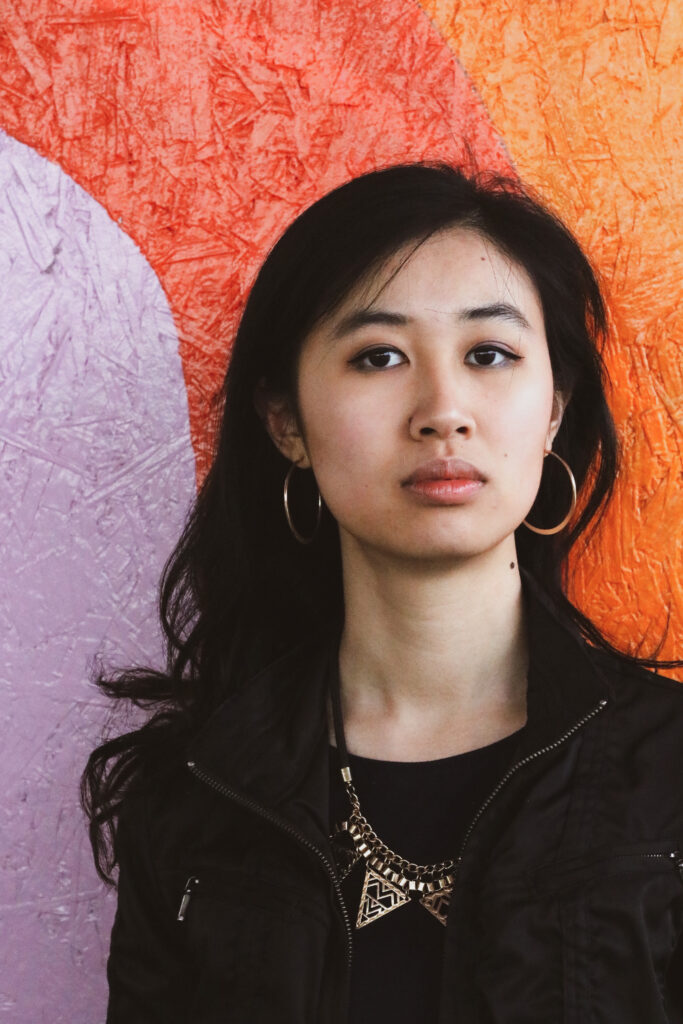 A young British East Asian woman with long black hair and large hoop earrings wearing a black zip-up jacket, standing in front of a colourful wall mural