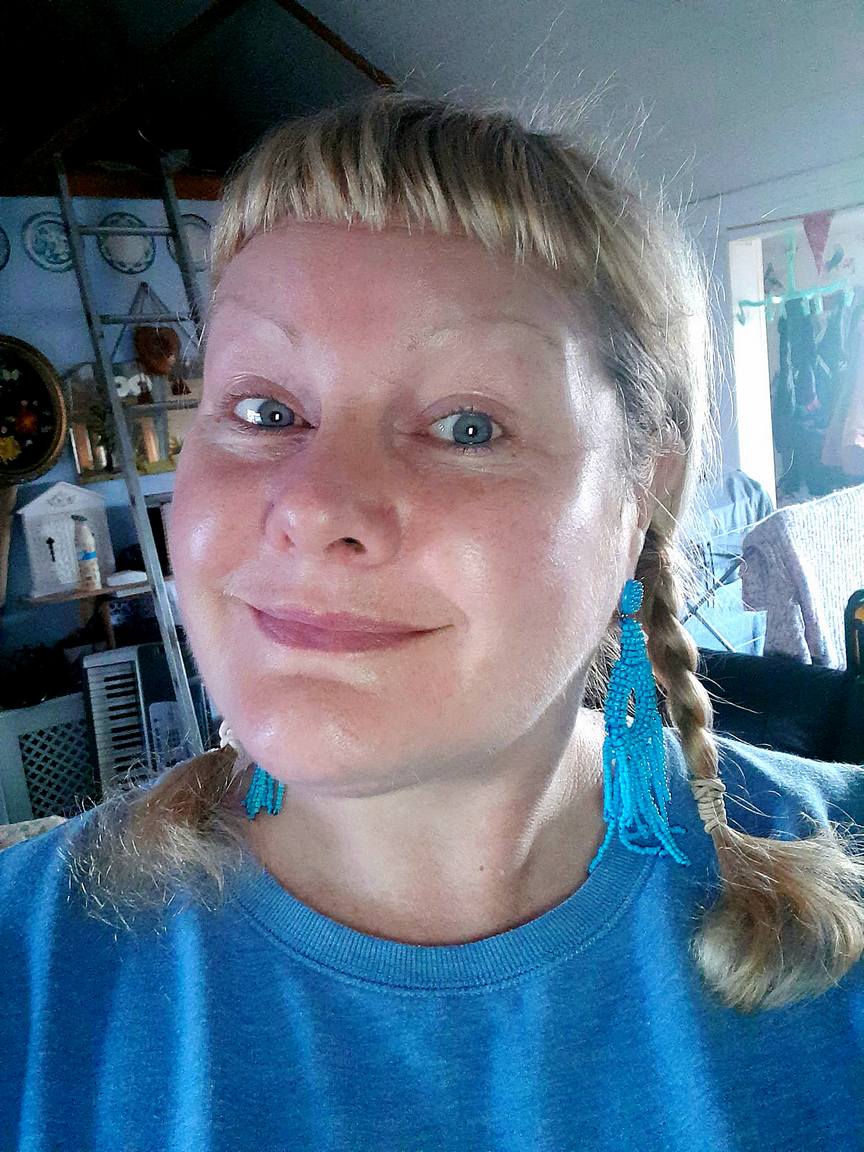A white woman in her middle years is smiling tightly at the camera and raising her eyebrows in exaggerated arches. She has blonde hair cut in a high, straight, 'Joan of Arc' fringe at the front, with pigtails either sie. She's wearing a bright turqoise blue round-necked top, and matching very long tasseled earrings.