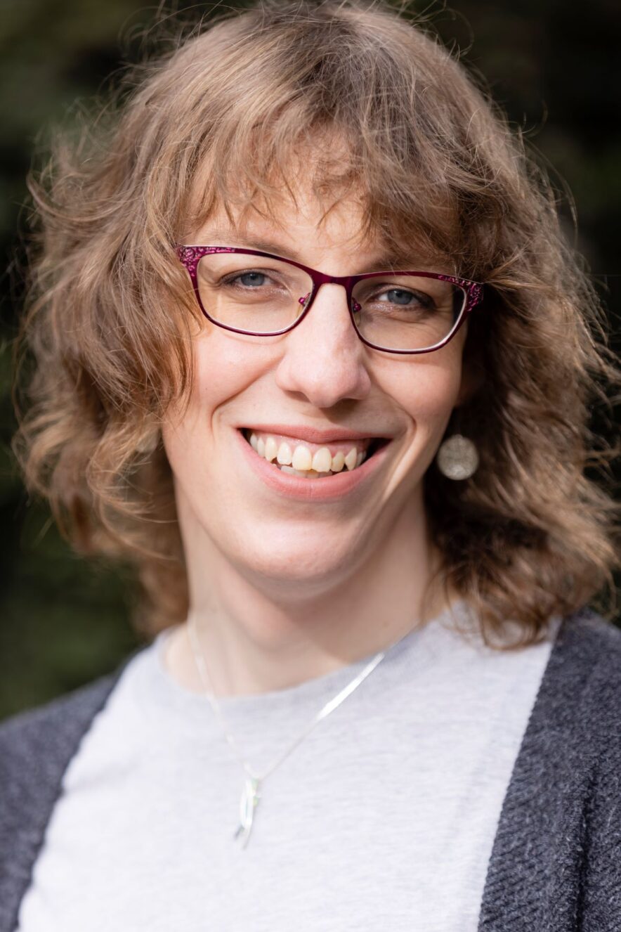 A white woman in her thirties with soft, waving, jaw-length light brown hair and red-framed squared off spectacles