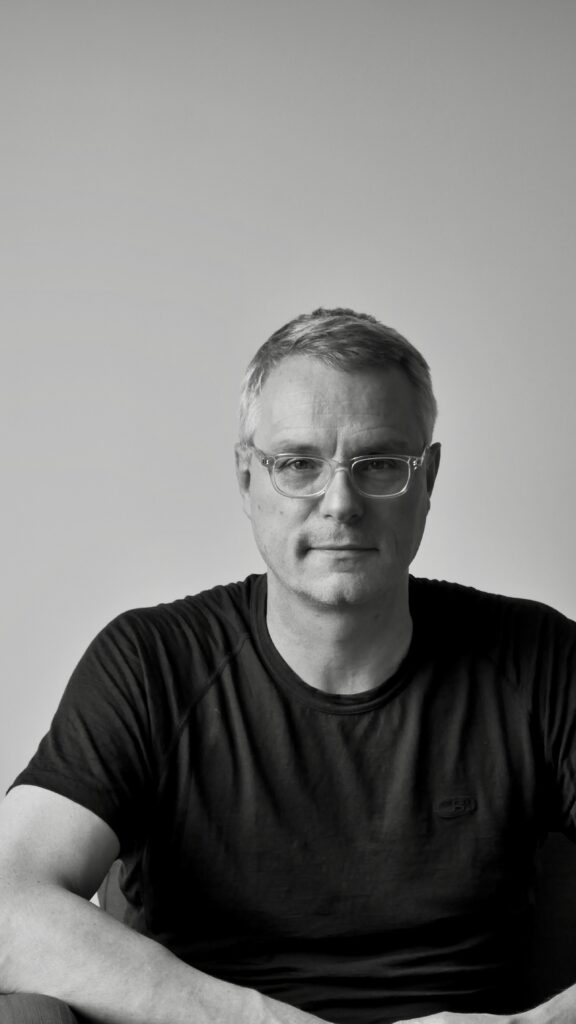 Black and white image of a white man with shor grey hair and clear-rimmed spectacles. wearing a dark short-sleeved t-shit. He looks relaxed and physically fit.