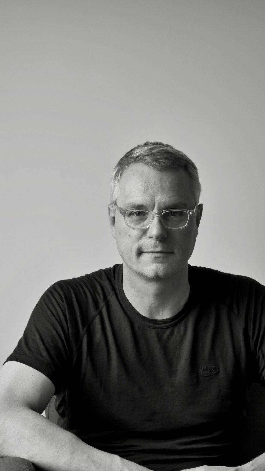 Black and white image of a white man with shor grey hair and clear-rimmed spectacles. wearing a dark short-sleeved t-shit. He looks relaxed and physically fit.