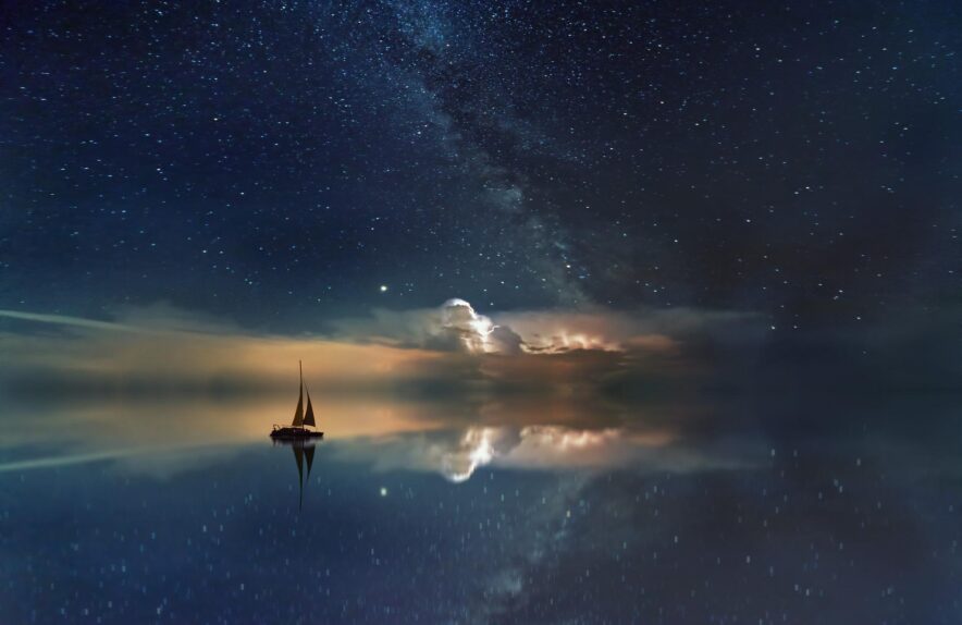 A small sailboat sets off into a dreamlike horizon. faintly orange-lit clouds and the Milky Way perfectly reflected in the still waters