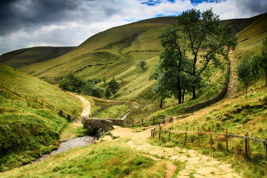 A sandy track through a small green valley, with a small stone bridge crossing a stream