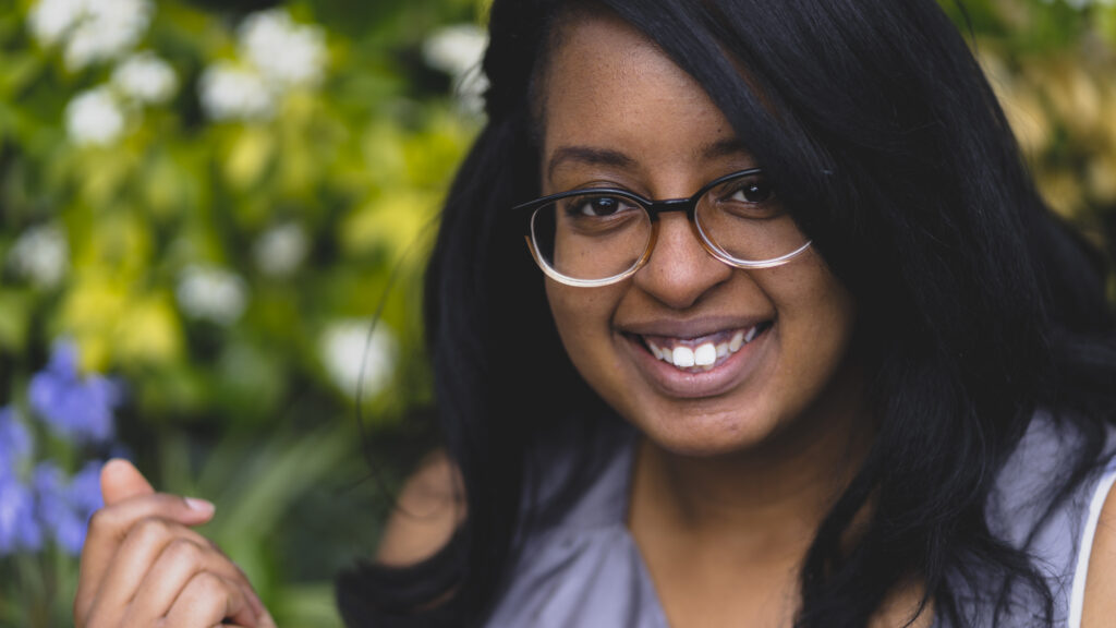 Jemilea Wisdom-Baako: A young Black woman with long, straight black hair, spectacles, and a happy, friendly smile