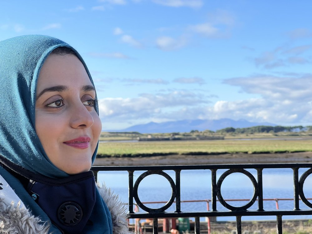 A brown woman in a teal blue hijab is standing in front of a wide landscape of river or estuary, with marshlands and distant mountains. The sky is a fresh spring-like blue.