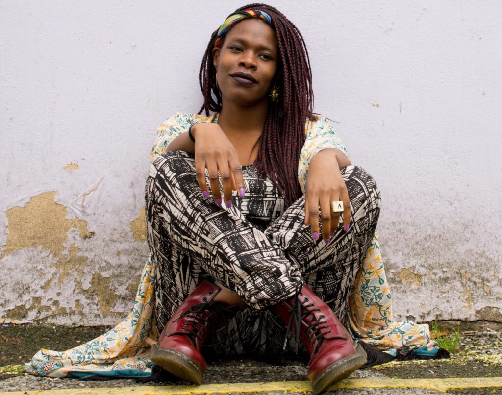 Young black woman with long plum-coloured braids and a wrapped fabric headband sits cross-legged against a wall of crumbling white plaster. She wears black and white print dungarees, a long yellow and blue kimono-wrap, and oxblood Doctor Martens. Her nails are violet, and one hand wears a chunky gold ring in the shape of an outsized Scrabble tile - 'V'